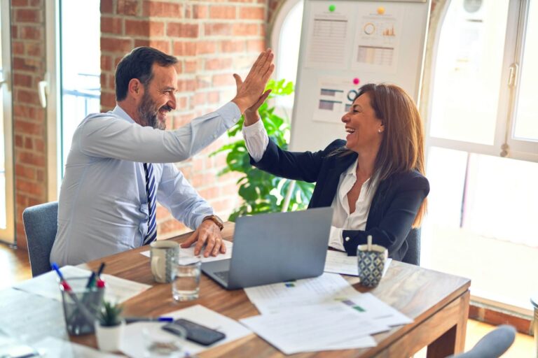 Business partners share a congratulatory high five while seated at a desk.